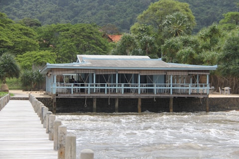 A wooden house on stilts built over the water, surrounded by lush green trees and vegetation. A wooden pier extends towards the house over the waves, which appear to be in motion. The structure features a covered porch with a simple railing and some furniture visible inside.