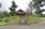 A rustic wooden house surrounded by lush greenery and vibrant sunflowers. The house has a triangular roof, and in front of it, there is a stone sign saying 'Kampot River Residence' amidst a small garden. Numerous trees and palm trees create a serene atmosphere.