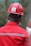 A person wearing a red hard hat and red protective workwear can be seen from behind. The helmet has a 'Safety First' sticker, emphasizing workplace safety. The person appears to be outdoors, and there is a green, blurred background, suggesting a construction or industrial setting.