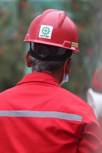 A person wearing a red hard hat and red protective workwear can be seen from behind. The helmet has a 'Safety First' sticker, emphasizing workplace safety. The person appears to be outdoors, and there is a green, blurred background, suggesting a construction or industrial setting.