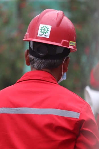 A person wearing a red hard hat and red protective workwear can be seen from behind. The helmet has a 'Safety First' sticker, emphasizing workplace safety. The person appears to be outdoors, and there is a green, blurred background, suggesting a construction or industrial setting.