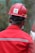 A person wearing a red hard hat and red protective workwear can be seen from behind. The helmet has a 'Safety First' sticker, emphasizing workplace safety. The person appears to be outdoors, and there is a green, blurred background, suggesting a construction or industrial setting.