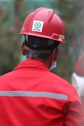 A person wearing a red hard hat and red protective workwear can be seen from behind. The helmet has a 'Safety First' sticker, emphasizing workplace safety. The person appears to be outdoors, and there is a green, blurred background, suggesting a construction or industrial setting.