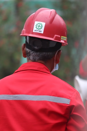 A person wearing a red hard hat and red protective workwear can be seen from behind. The helmet has a 'Safety First' sticker, emphasizing workplace safety. The person appears to be outdoors, and there is a green, blurred background, suggesting a construction or industrial setting.