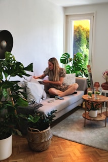A woman is sitting on a modern gray sofa in a bright living room, surrounded by green plants. She holds a pencil or pen, writing or drawing on a notepad resting on a pillow on her lap. A small wooden side table nearby holds two glasses of green beverage. A guitar is propped against the wall, complementing the cozy and artistic atmosphere, and sunlight filters through a window with a view of lush greenery outside.