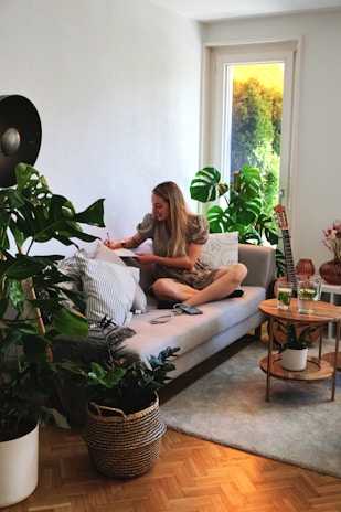 A woman is sitting on a modern gray sofa in a bright living room, surrounded by green plants. She holds a pencil or pen, writing or drawing on a notepad resting on a pillow on her lap. A small wooden side table nearby holds two glasses of green beverage. A guitar is propped against the wall, complementing the cozy and artistic atmosphere, and sunlight filters through a window with a view of lush greenery outside.
