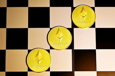 Three gold coins with the Ethereum logo and text are placed diagonally on a black and white checkerboard surface.