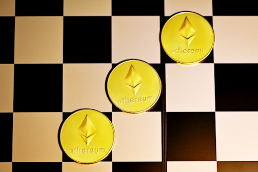 Three gold coins with the Ethereum logo and text are placed diagonally on a black and white checkerboard surface.