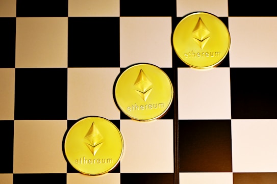 Three gold coins with the Ethereum logo and text are placed diagonally on a black and white checkerboard surface.