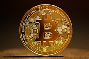 A highly detailed, golden Bitcoin token stands vertically against a dark, blurred background. The intricate design features circuitry patterns on the surface with the Bitcoin logo prominently displayed in the center. Engravings around the coin's edges include words like 'Blockchain', 'Peer to Peer', and 'Decentralized'. The lighting highlights the metallic sheen of the coin, casting a soft shadow on the surface below.