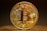 A highly detailed, golden Bitcoin token stands vertically against a dark, blurred background. The intricate design features circuitry patterns on the surface with the Bitcoin logo prominently displayed in the center. Engravings around the coin's edges include words like 'Blockchain', 'Peer to Peer', and 'Decentralized'. The lighting highlights the metallic sheen of the coin, casting a soft shadow on the surface below.
