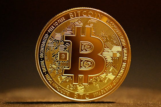 A highly detailed, golden Bitcoin token stands vertically against a dark, blurred background. The intricate design features circuitry patterns on the surface with the Bitcoin logo prominently displayed in the center. Engravings around the coin's edges include words like 'Blockchain', 'Peer to Peer', and 'Decentralized'. The lighting highlights the metallic sheen of the coin, casting a soft shadow on the surface below.
