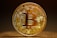 A highly detailed, golden Bitcoin token stands vertically against a dark, blurred background. The intricate design features circuitry patterns on the surface with the Bitcoin logo prominently displayed in the center. Engravings around the coin's edges include words like 'Blockchain', 'Peer to Peer', and 'Decentralized'. The lighting highlights the metallic sheen of the coin, casting a soft shadow on the surface below.