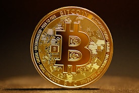 A highly detailed, golden Bitcoin token stands vertically against a dark, blurred background. The intricate design features circuitry patterns on the surface with the Bitcoin logo prominently displayed in the center. Engravings around the coin's edges include words like 'Blockchain', 'Peer to Peer', and 'Decentralized'. The lighting highlights the metallic sheen of the coin, casting a soft shadow on the surface below.