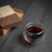 A block of compressed tea is partially unwrapped in brown paper, placed on a black wooden surface. Next to it is a transparent glass cup filled with dark tea. The setting is minimalistic and elegant, highlighting the textures of the tea block and the wood grain.