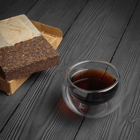 A block of compressed tea is partially unwrapped in brown paper, placed on a black wooden surface. Next to it is a transparent glass cup filled with dark tea. The setting is minimalistic and elegant, highlighting the textures of the tea block and the wood grain.