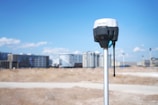 A surveying instrument mounted on a tall pole stands in the foreground, with a bright blue sky and scattered clouds above. In the background, there are several modern high-rise buildings, indicating an urban area.