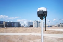 A surveying instrument mounted on a tall pole stands in the foreground, with a bright blue sky and scattered clouds above. In the background, there are several modern high-rise buildings, indicating an urban area.