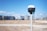 A surveying instrument mounted on a tall pole stands in the foreground, with a bright blue sky and scattered clouds above. In the background, there are several modern high-rise buildings, indicating an urban area.