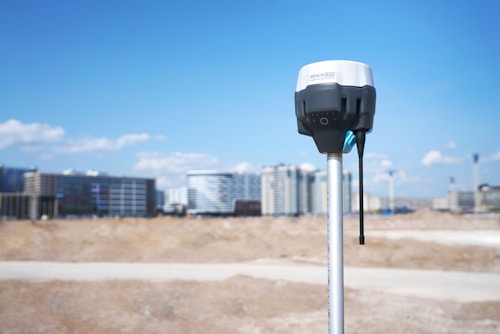 A surveying instrument mounted on a tall pole stands in the foreground, with a bright blue sky and scattered clouds above. In the background, there are several modern high-rise buildings, indicating an urban area.