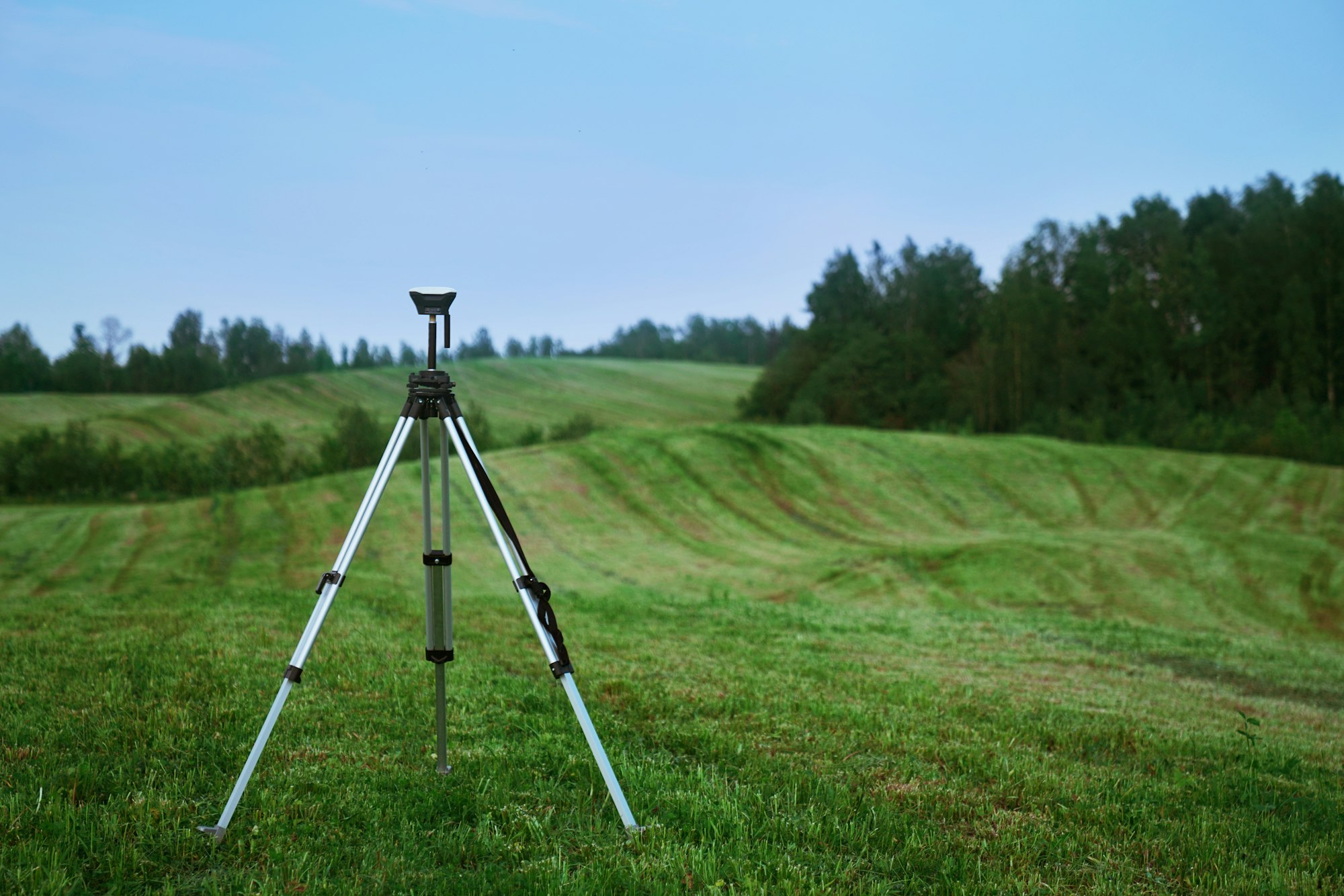 A surveyors device on a tripod in a lush green field of rolling hills, trees in the background, at dusk.