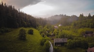 A serene countryside landscape featuring lush green hills and dense forests under a cloudy sky. Several rustic houses are nestled among the trees. Mist hovers above the trees in the distance, adding a touch of mystery to the scene.