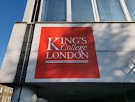 A large red signboard with white text displaying 'King's College London' is mounted on a building with white brickwork. The building features large windows above the sign, reflecting a clear blue sky.