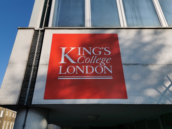 A large red signboard with white text displaying 'King's College London' is mounted on a building with white brickwork. The building features large windows above the sign, reflecting a clear blue sky.