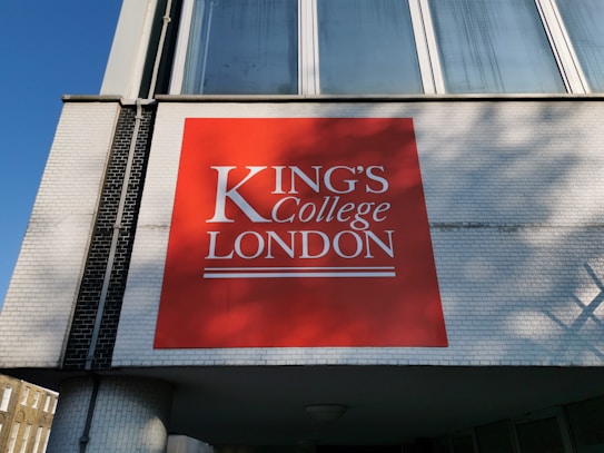 A large red signboard with white text displaying 'King's College London' is mounted on a building with white brickwork. The building features large windows above the sign, reflecting a clear blue sky.