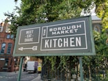 A large green sign with white text is positioned near a lush green tree. The sign reads 'Meet Eat Share' on the left and 'The Borough Market Kitchen' on the right. An arrow pointing left is also visible. In the background, there is a street with a white truck and buildings made of brick with arched windows.