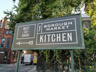 A large green sign with white text is positioned near a lush green tree. The sign reads 'Meet Eat Share' on the left and 'The Borough Market Kitchen' on the right. An arrow pointing left is also visible. In the background, there is a street with a white truck and buildings made of brick with arched windows.