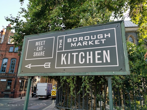 A large green sign with white text is positioned near a lush green tree. The sign reads 'Meet Eat Share' on the left and 'The Borough Market Kitchen' on the right. An arrow pointing left is also visible. In the background, there is a street with a white truck and buildings made of brick with arched windows.