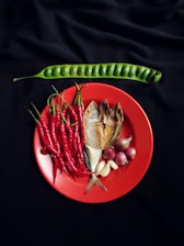 A vibrant arrangement of fresh ingredients is displayed on a red plate, including red chili peppers, smoked fish, garlic, and shallots. Positioned above on a black background is a long, green bean pod.