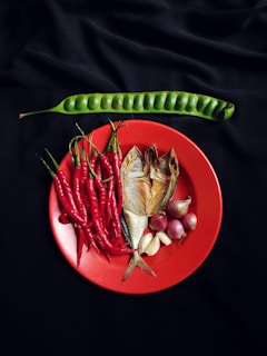 A vibrant arrangement of fresh ingredients is displayed on a red plate, including red chili peppers, smoked fish, garlic, and shallots. Positioned above on a black background is a long, green bean pod.