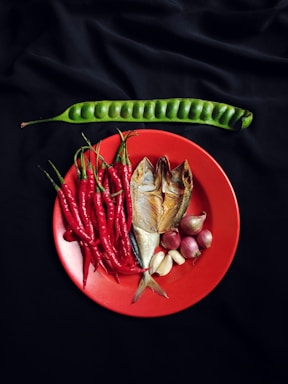 A vibrant arrangement of fresh ingredients is displayed on a red plate, including red chili peppers, smoked fish, garlic, and shallots. Positioned above on a black background is a long, green bean pod.