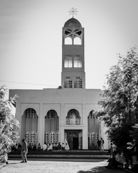 A tall church building with a prominent steeple and cross on top stands against the sky. The façade features arched windows and a central entrance. People are gathered near the entrance, with some sitting on the steps.