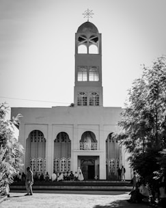A tall church building with a prominent steeple and cross on top stands against the sky. The façade features arched windows and a central entrance. People are gathered near the entrance, with some sitting on the steps.