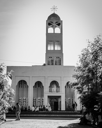 A tall church building with a prominent steeple and cross on top stands against the sky. The façade features arched windows and a central entrance. People are gathered near the entrance, with some sitting on the steps.
