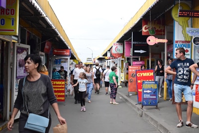 A busy outdoor marketplace with people walking between rows of small shops and colorful signage in various languages. The stands have a variety of posters and advertisements, and the atmosphere is lively. Individuals of different ages and styles are moving around, some carrying bags.