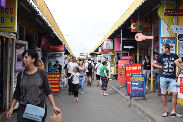 A busy outdoor marketplace with people walking between rows of small shops and colorful signage in various languages. The stands have a variety of posters and advertisements, and the atmosphere is lively. Individuals of different ages and styles are moving around, some carrying bags.