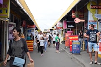 A busy outdoor marketplace with people walking between rows of small shops and colorful signage in various languages. The stands have a variety of posters and advertisements, and the atmosphere is lively. Individuals of different ages and styles are moving around, some carrying bags.