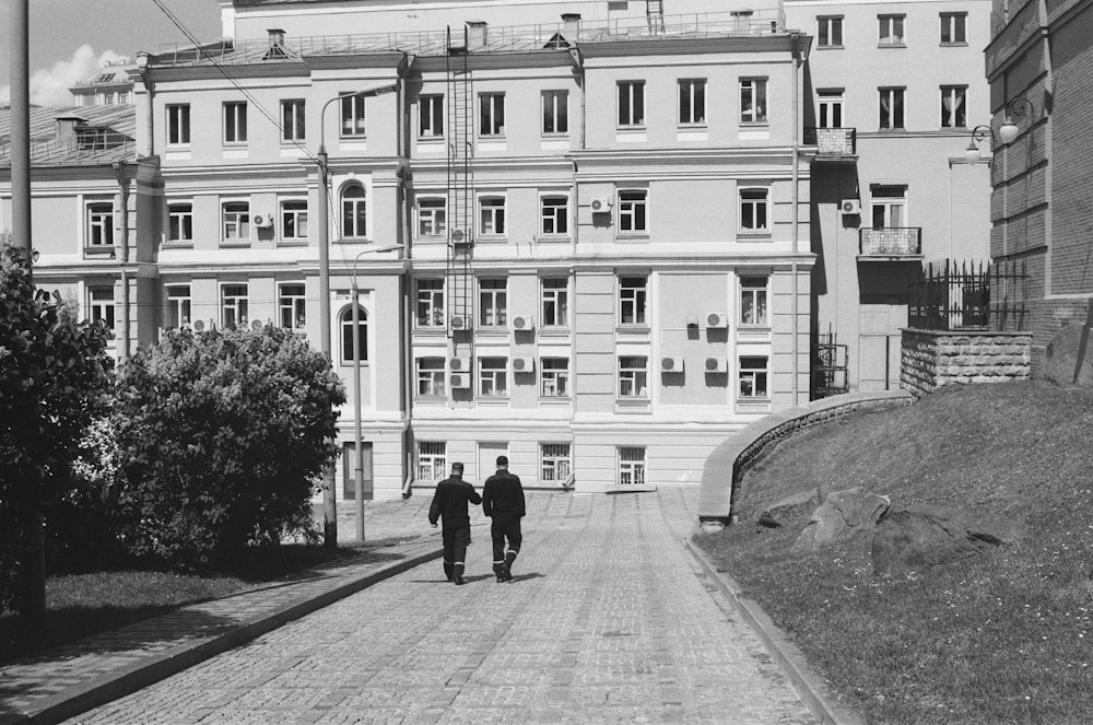 grayscale photo of 2 person walking on sidewalk near building