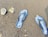A pair of blue flip-flops with a checkered pattern and the word 'Quiksilver' printed on them, placed on dark, coarse sand. Nearby, there are a couple of small rocks or pebbles partially embedded in the sand.