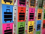 A row of colorful lockers with luggage symbols and numbers on their doors. Each locker is brightly colored in shades such as blue, pink, green, yellow, and orange, and features a large, black suitcase icon. Some lockers have opened red locks hanging on them.