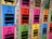 A row of colorful lockers with luggage symbols and numbers on their doors. Each locker is brightly colored in shades such as blue, pink, green, yellow, and orange, and features a large, black suitcase icon. Some lockers have opened red locks hanging on them.