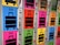 A row of colorful lockers with luggage symbols and numbers on their doors. Each locker is brightly colored in shades such as blue, pink, green, yellow, and orange, and features a large, black suitcase icon. Some lockers have opened red locks hanging on them.