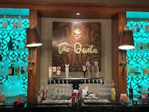 A bar setup featuring a decorative wooden sign with the word 'Tequila' in the center, surrounded by intricate carvings. Shelves on either side are filled with various bottles of liquor, each illuminated by a turquoise backlight. Below the sign, several beer taps are lined up along with an assortment of glassware and drink mixers.