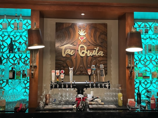 A bar setup featuring a decorative wooden sign with the word 'Tequila' in the center, surrounded by intricate carvings. Shelves on either side are filled with various bottles of liquor, each illuminated by a turquoise backlight. Below the sign, several beer taps are lined up along with an assortment of glassware and drink mixers.