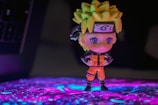 A small figurine of an anime character stands confidently with arms crossed, wearing an orange jumpsuit and a headband with a unique symbol. The background features a colorful, illuminated surface with a blend of purple and blue hues.