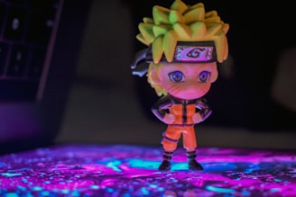 A small figurine of an anime character stands confidently with arms crossed, wearing an orange jumpsuit and a headband with a unique symbol. The background features a colorful, illuminated surface with a blend of purple and blue hues.